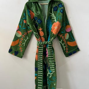 May include: A long-sleeved green velvet robe featuring a vibrant floral embroidery pattern in shades of orange, pink, blue, and light green. The garment includes a matching tie belt and a collar, with a striped lining visible at the neck and belt.