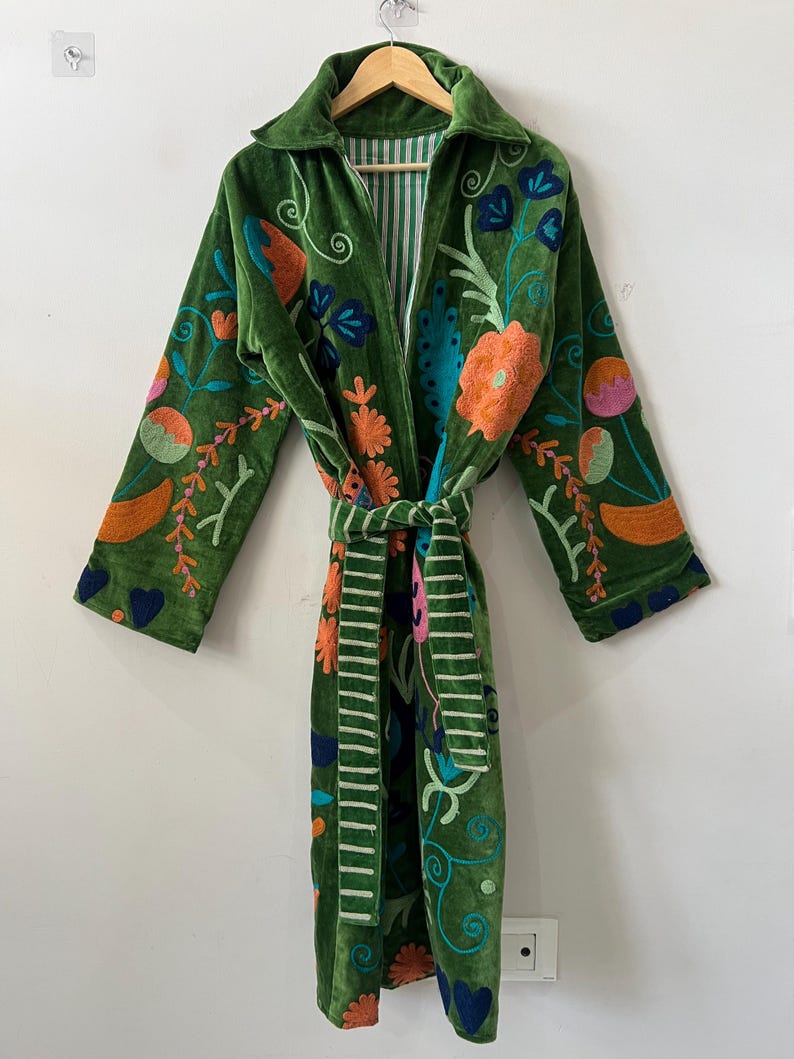 May include: A long-sleeved green velvet robe featuring a vibrant floral embroidery pattern in shades of orange, pink, blue, and light green. The garment includes a matching tie belt and a collar, with a striped lining visible at the neck and belt.