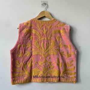 May include: A sleeveless pink vest with intricate gold embroidery. The embroidery features a floral design that covers the front and back of the vest. The vest is hanging on a wooden hanger.