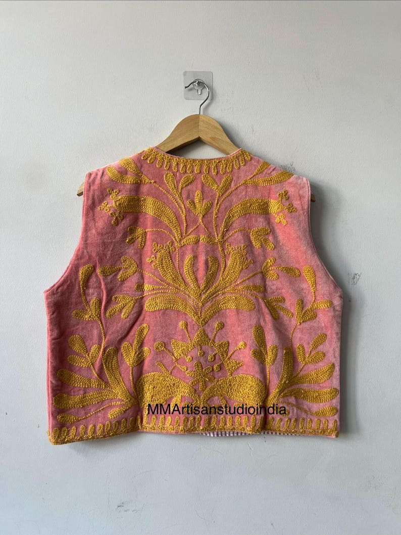 May include: A sleeveless pink vest with intricate gold embroidery. The embroidery features a floral design that covers the front and back of the vest. The vest is hanging on a wooden hanger.