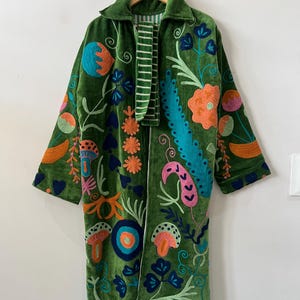 May include: A long-sleeved green velvet coat featuring vibrant, multi-colored embroidered botanical and mushroom motifs. The garment includes a collar, an open front, and a striped fabric lining visible at the neckline and along the inner edges.