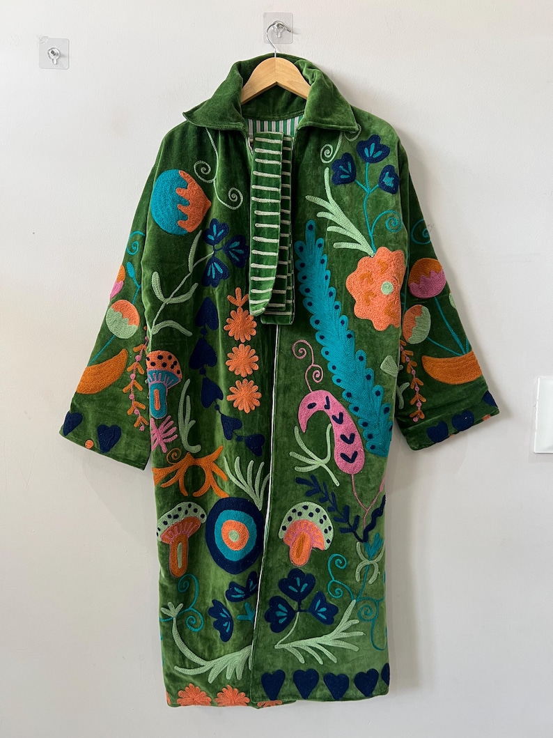 May include: A long-sleeved green velvet coat featuring vibrant, multi-colored embroidered botanical and mushroom motifs. The garment includes a collar, an open front, and a striped fabric lining visible at the neckline and along the inner edges.