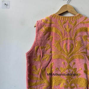 May include: A sleeveless pink vest with intricate gold embroidery. The vest features a floral design and is hanging on a wooden hanger. The text "MMArtisanstudioindia" is visible on the vest.