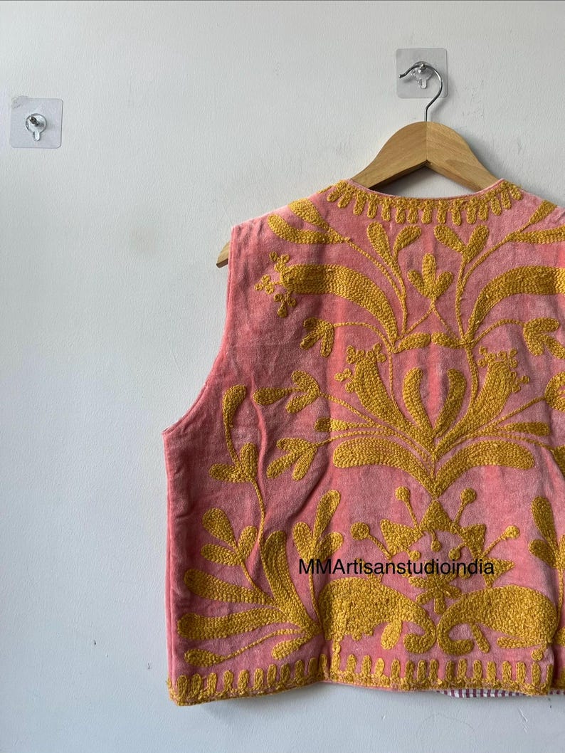 May include: A sleeveless pink vest with intricate gold embroidery. The vest features a floral design and is hanging on a wooden hanger. The text "MMArtisanstudioindia" is visible on the vest.