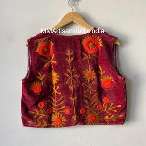 Puede incluir: Chaleco burdeos con bordados florales en naranja y dorado, colgado de una percha de madera. El texto "MMArtisanstudioindia" es visible. Prenda de vestir decorativa.