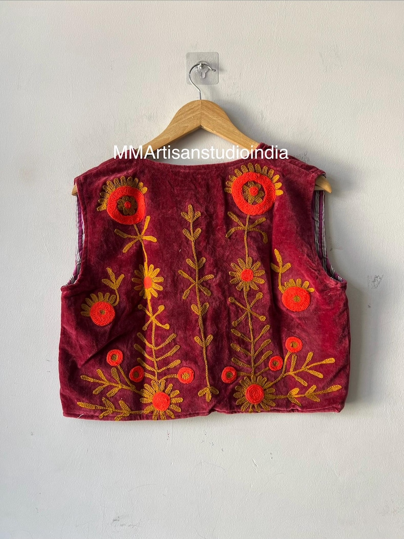 Puede incluir: Chaleco burdeos con bordados florales en naranja y dorado, colgado de una percha de madera. El texto "MMArtisanstudioindia" es visible. Prenda de vestir decorativa.