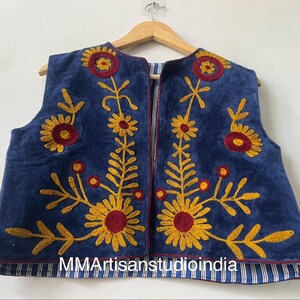 May include: A sleeveless blue vest with embroidered yellow and maroon floral designs. The vest is hanging on a wooden hanger. The bottom hem is lined with blue and white striped fabric. The text "MMArtisanstudioindia" is visible at the bottom.