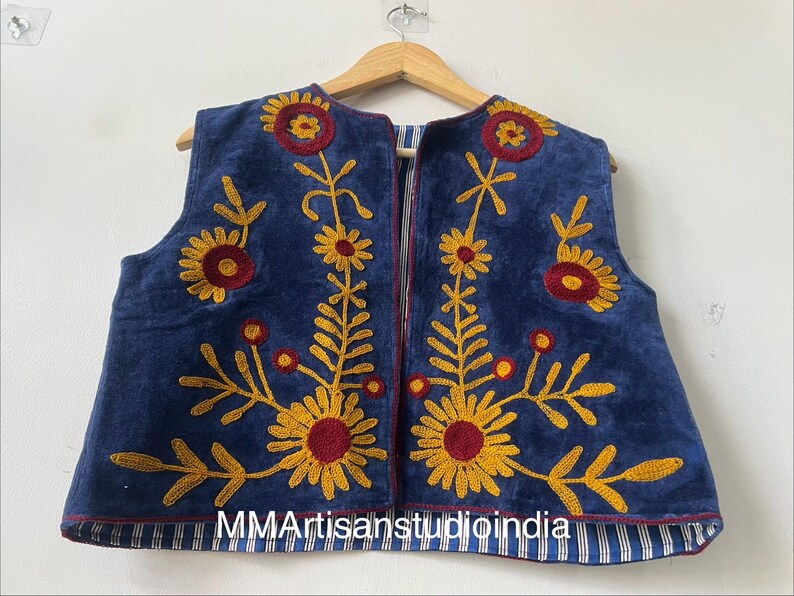 May include: A sleeveless blue vest with embroidered yellow and maroon floral designs. The vest is hanging on a wooden hanger. The bottom hem is lined with blue and white striped fabric. The text "MMArtisanstudioindia" is visible at the bottom.