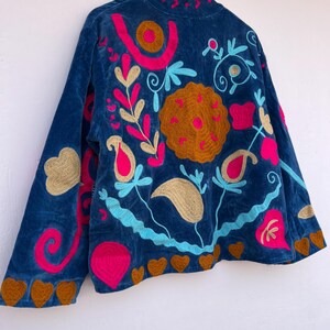 May include: A blue velvet jacket with colorful floral embroidery. The jacket features a high collar and long sleeves. The embroidery includes pink, gold, and blue floral designs, with brown heart-shaped accents along the hem and sleeve cuffs.