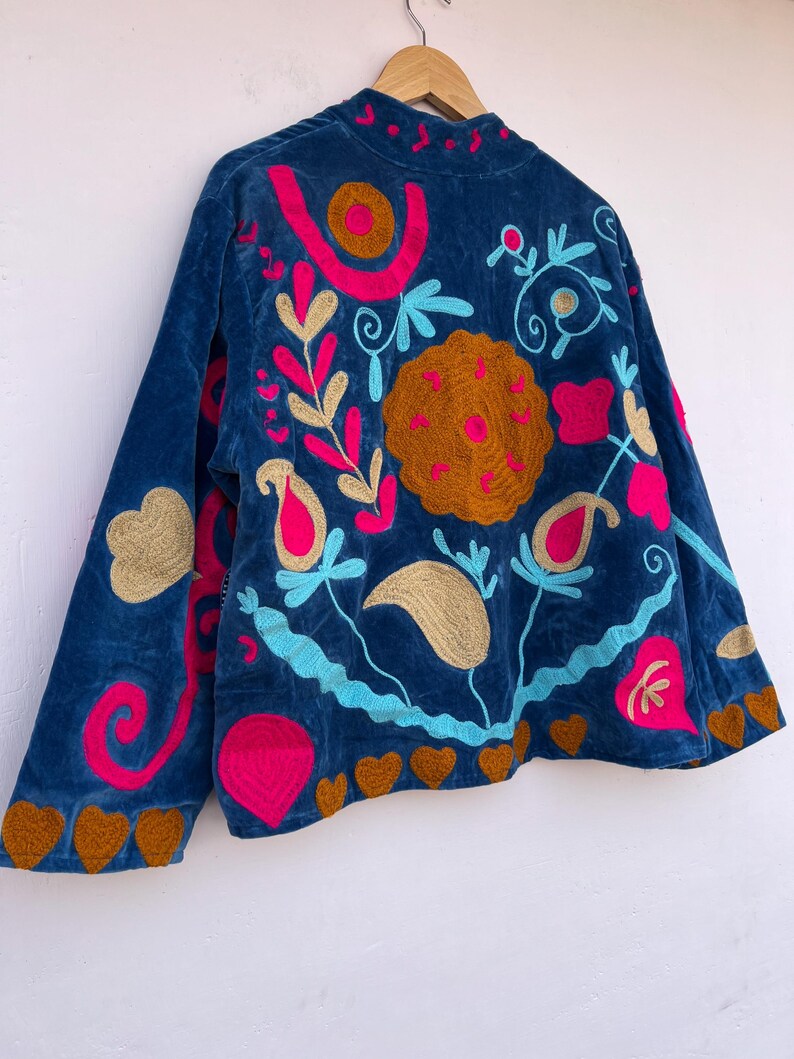 May include: A blue velvet jacket with colorful floral embroidery. The jacket features a high collar and long sleeves. The embroidery includes pink, gold, and blue floral designs, with brown heart-shaped accents along the hem and sleeve cuffs.