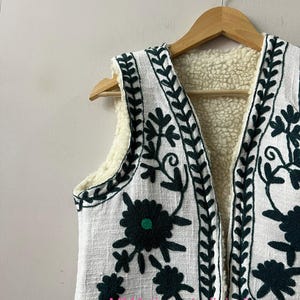 May include: White vest with dark green floral embroidery and a cream-colored fleece lining. The vest is hanging on a wooden hanger. The embroidery includes floral and leaf patterns.