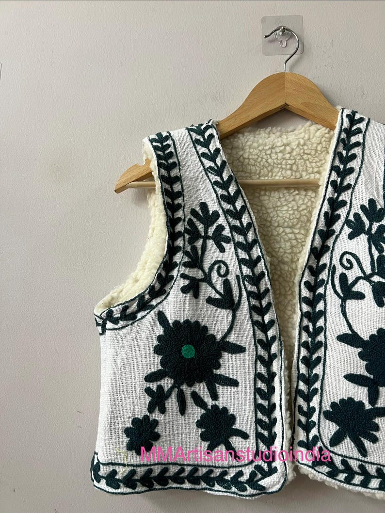 May include: White vest with dark green floral embroidery and a cream-colored fleece lining. The vest is hanging on a wooden hanger. The embroidery includes floral and leaf patterns.