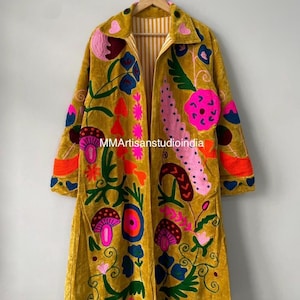 May include: A mustard yellow velvet long coat featuring vibrant multi-colored floral and mushroom embroidery. The garment has a collar, long sleeves, and an open front with a white and yellow striped lining. The text MMArtisanstudioindia is visible.