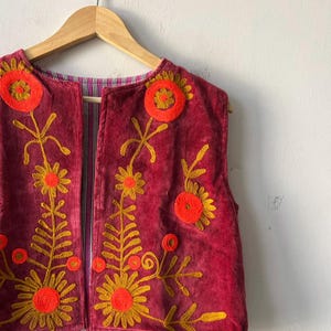 Puede incluir: Chaleco burdeos sin mangas con dise&ntilde;os florales bordados en dorado y naranja. El chaleco est&aacute; colgado de una percha de madera. El forro interior presenta un estampado a rayas.
