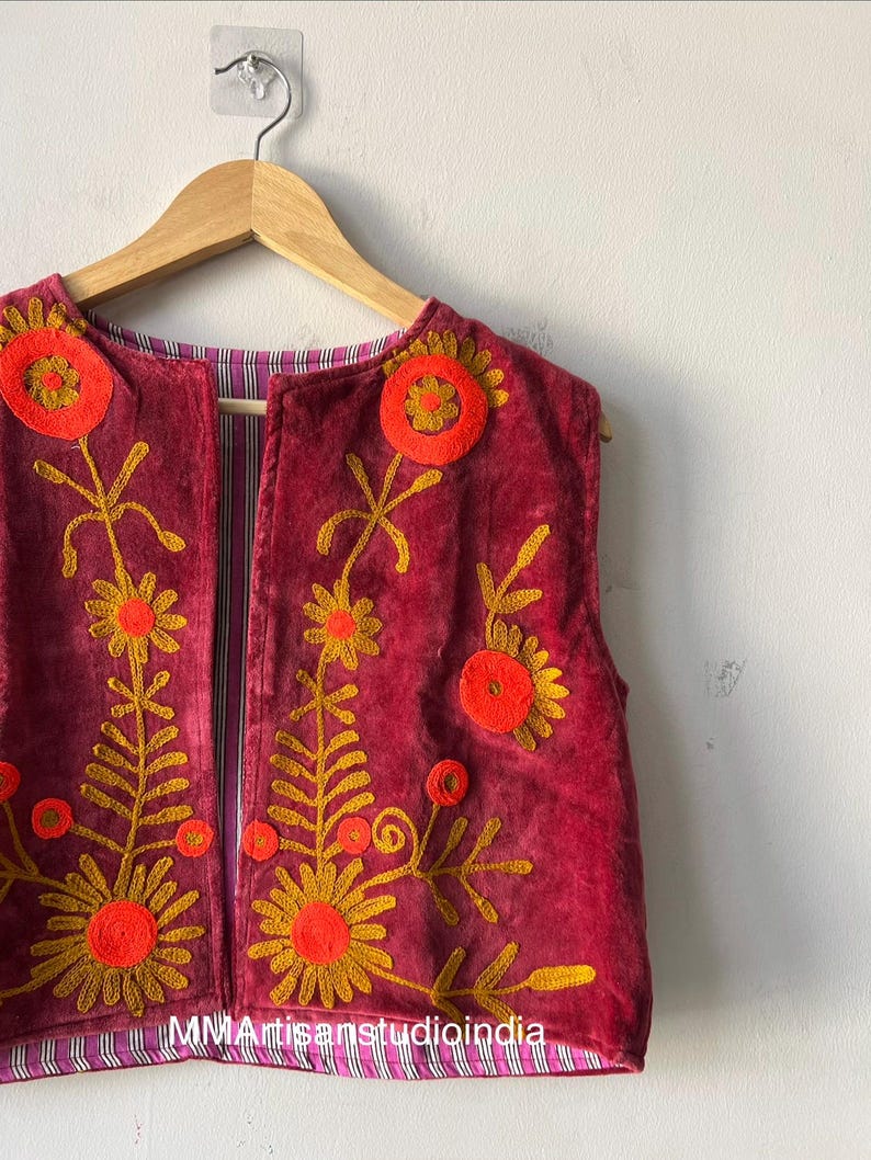 Puede incluir: Chaleco burdeos sin mangas con dise&ntilde;os florales bordados en dorado y naranja. El chaleco est&aacute; colgado de una percha de madera. El forro interior presenta un estampado a rayas.