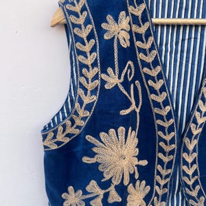 May include: A blue embroidered vest with a beige floral design. The vest is made of a soft, velvety material and has a striped lining. The vest is hanging on a wooden hanger.