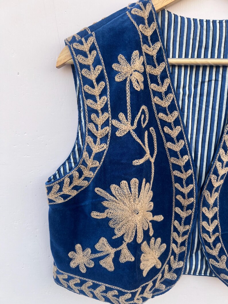 May include: A blue embroidered vest with a beige floral design. The vest is made of a soft, velvety material and has a striped lining. The vest is hanging on a wooden hanger.