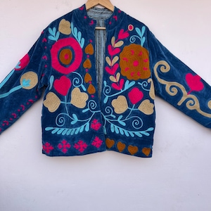 May include: A blue velvet jacket with colorful floral embroidery. The jacket features pink, gold, and turquoise embroidered flowers and swirls. The jacket has a striped lining and a collarless design.