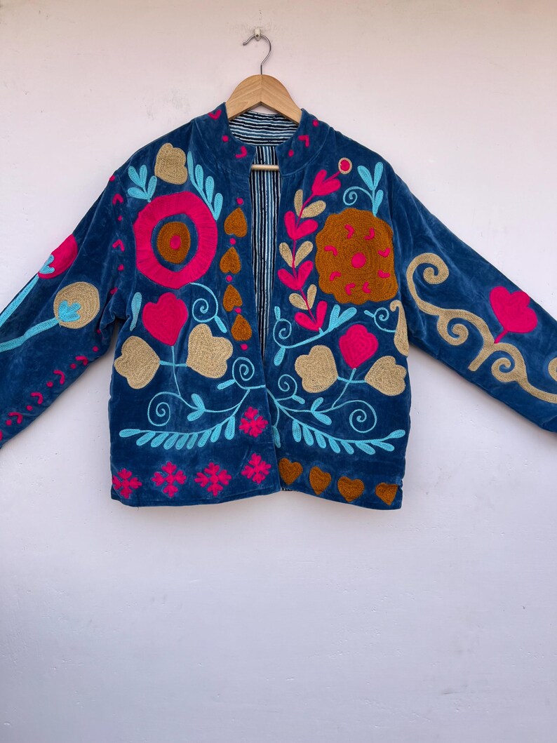 May include: A blue velvet jacket with colorful floral embroidery. The jacket features pink, gold, and turquoise embroidered flowers and swirls. The jacket has a striped lining and a collarless design.