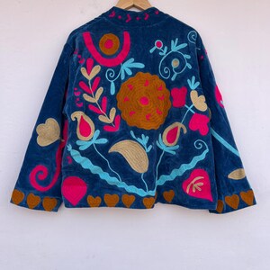 May include: A dark blue velvet jacket with colorful floral embroidery. The jacket features a high collar and long sleeves. The embroidery includes pink, gold, and turquoise designs, with a brown heart-shaped trim along the bottom.