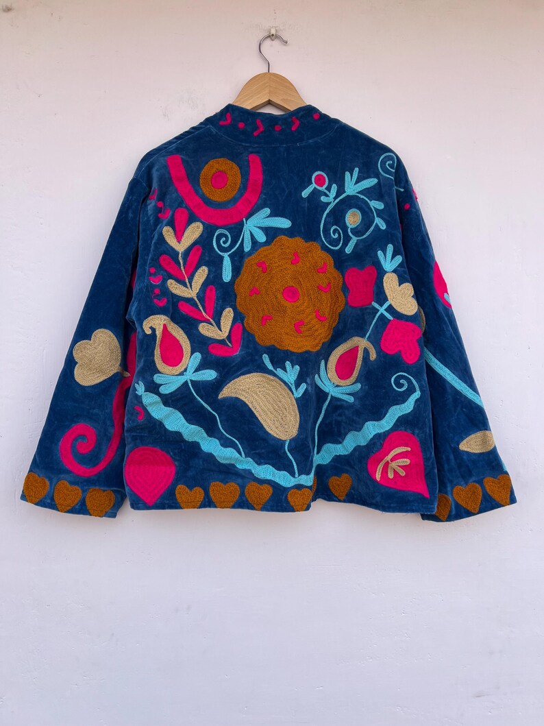 May include: A dark blue velvet jacket with colorful floral embroidery. The jacket features a high collar and long sleeves. The embroidery includes pink, gold, and turquoise designs, with a brown heart-shaped trim along the bottom.