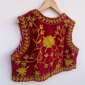 May include: A red velvet vest with gold floral embroidery, hanging on a wooden hanger. The sleeveless garment features a detailed design of flowers and leaves. The background is plain white.