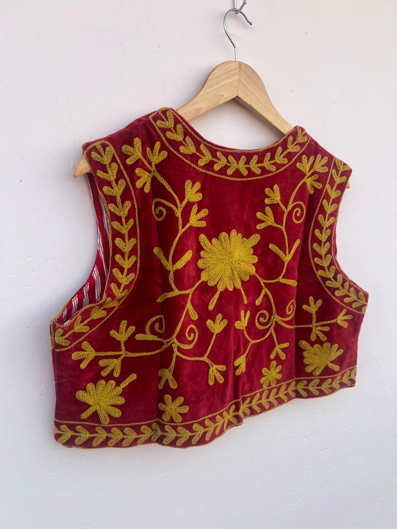 May include: A red velvet vest with gold floral embroidery, hanging on a wooden hanger. The sleeveless garment features a detailed design of flowers and leaves. The background is plain white.