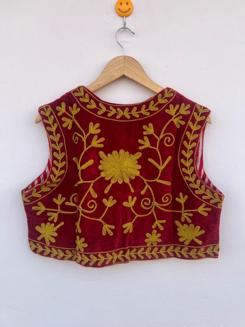 May include: A red velvet vest with gold floral embroidery, displayed on a wooden hanger. The embroidery includes a central flower and detailed vine patterns. The sleeveless vest is against a plain white background. The garment is a traditional style.