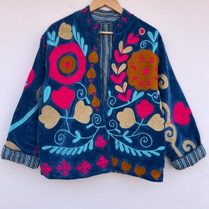 May include: A blue velvet jacket with floral embroidery in pink, gold, and turquoise. The jacket features a striped lining and cuffs. The design includes large flower and leaf patterns.