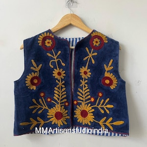 May include: A sleeveless, navy blue vest with embroidered floral designs in yellow and red. The vest has a striped lining and is displayed on a wooden hanger. The text "MMArtisanstudioindia" is visible on the bottom.