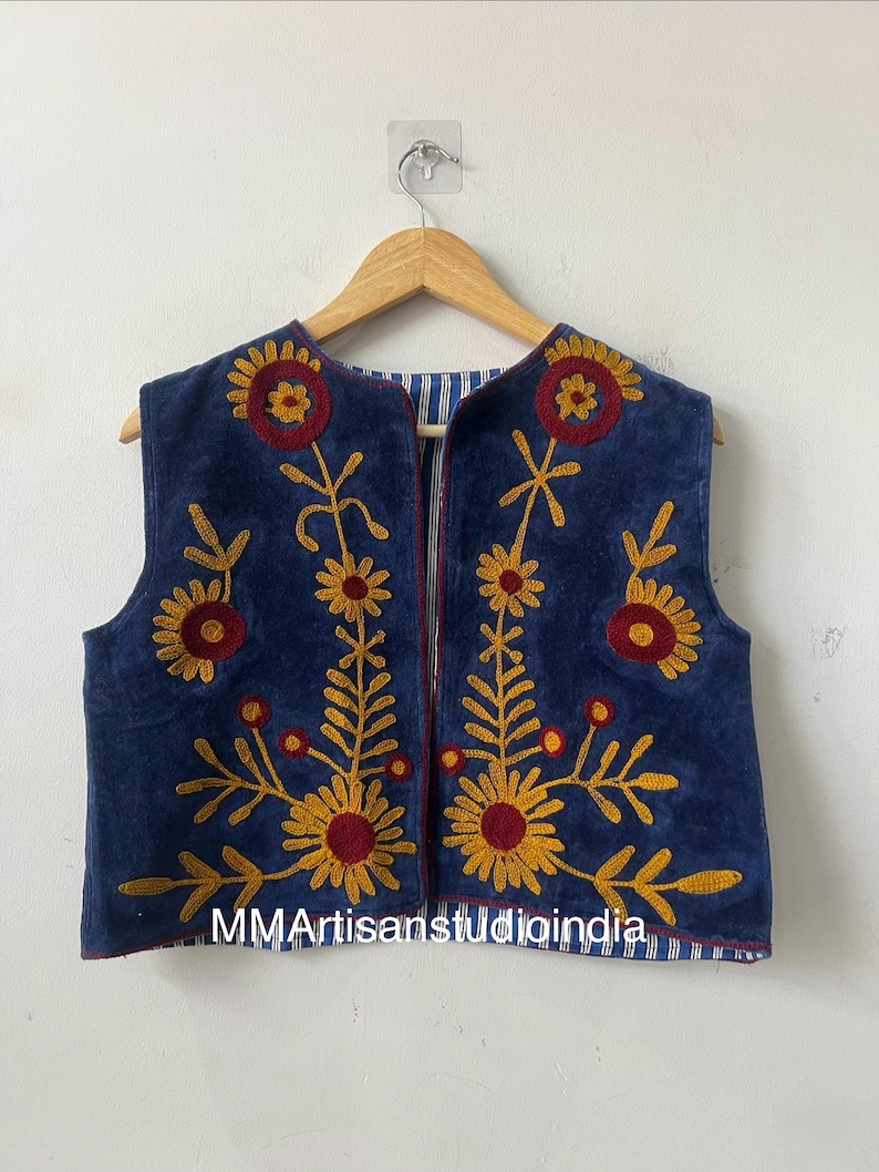 May include: A sleeveless, navy blue vest with embroidered floral designs in yellow and red. The vest has a striped lining and is displayed on a wooden hanger. The text "MMArtisanstudioindia" is visible on the bottom.