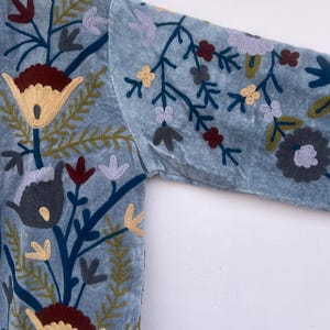 以下が含まれることがあります： 刺繍の花柄が施された、ライトブルーのベルベットジャケット。刺繍は、クリーム色、バーガンディ色、黄色、グレーの花と、緑と青の茎と葉が特徴です。ジャケットの袖が見え、複雑な花柄が際立っています。