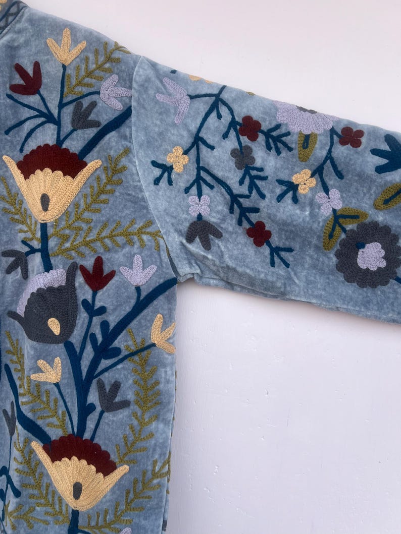 以下が含まれることがあります： 刺繍の花柄が施された、ライトブルーのベルベットジャケット。刺繍は、クリーム色、バーガンディ色、黄色、グレーの花と、緑と青の茎と葉が特徴です。ジャケットの袖が見え、複雑な花柄が際立っています。