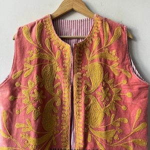 May include: A sleeveless, open-front vest in a coral hue, adorned with intricate yellow embroidery. The embroidery features floral and paisley-like designs. The vest's interior lining showcases a red and white striped pattern. The garment is displayed on a wooden hanger.