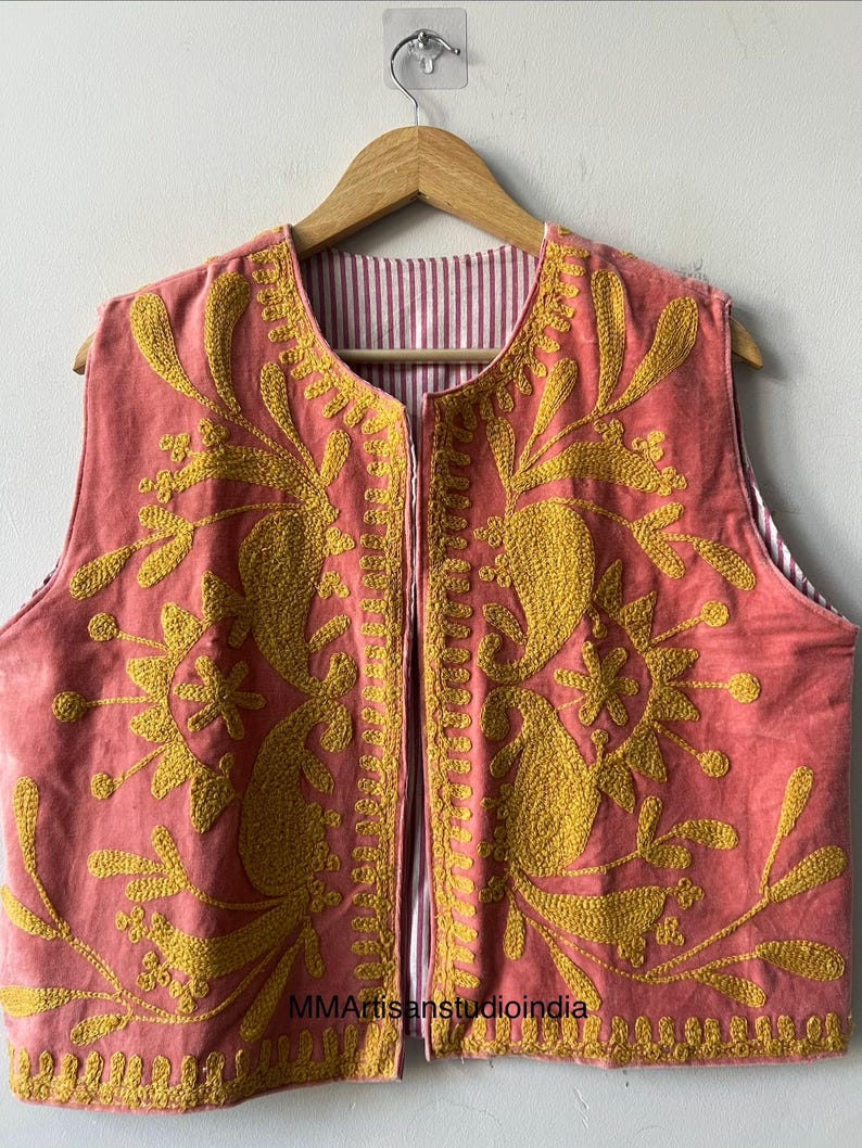 May include: A sleeveless, open-front vest in a coral hue, adorned with intricate yellow embroidery. The embroidery features floral and paisley-like designs. The vest's interior lining showcases a red and white striped pattern. The garment is displayed on a wooden hanger.