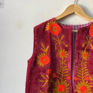 Puede incluir: Un chaleco burdeos sin mangas con dise&ntilde;os florales bordados en naranja y dorado. El chaleco est&aacute; colgado en una percha de madera contra una pared blanca. El forro interior presenta un patr&oacute;n de rayas. El texto "MMArtisanstudioindia" es visible.