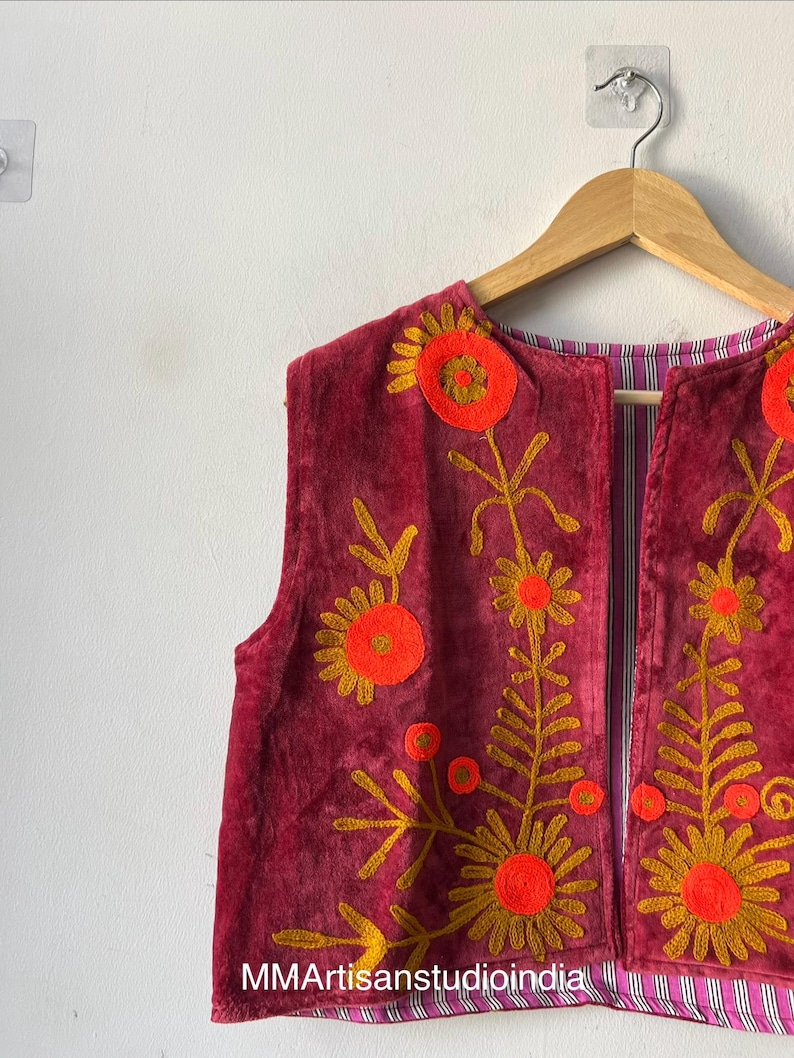 Puede incluir: Un chaleco burdeos sin mangas con dise&ntilde;os florales bordados en naranja y dorado. El chaleco est&aacute; colgado en una percha de madera contra una pared blanca. El forro interior presenta un patr&oacute;n de rayas. El texto "MMArtisanstudioindia" es visible.