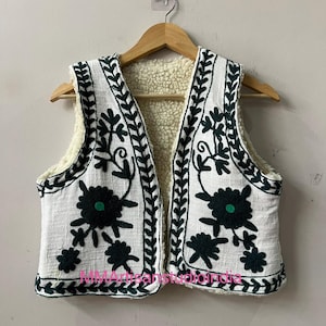 May include: White vest with dark green floral embroidery and a cream-colored fleece lining. The sleeveless vest is hanging on a wooden hanger. The vest has a textured fabric and a decorative border.