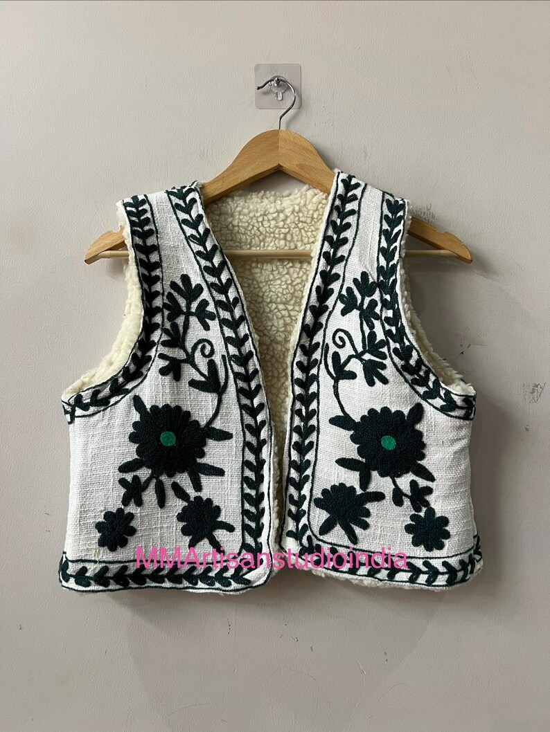 May include: White vest with dark green floral embroidery and a cream-colored fleece lining. The sleeveless vest is hanging on a wooden hanger. The vest has a textured fabric and a decorative border.