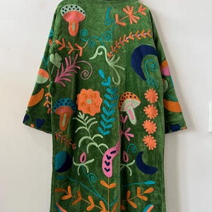 May include: A long-sleeved green coat featuring vibrant, multi-colored embroidery. The design includes various botanical and celestial motifs such as mushrooms, flowers, leaves, and crescent moons in shades of orange, pink, blue, and teal.