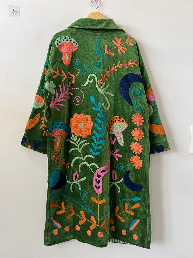 May include: A long-sleeved green coat featuring vibrant, multi-colored embroidery. The design includes various botanical and celestial motifs such as mushrooms, flowers, leaves, and crescent moons in shades of orange, pink, blue, and teal.