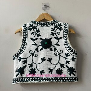 May include: White vest with intricate dark green floral embroidery. The sleeveless garment features a cream-colored sherpa lining and is displayed on a wooden hanger. The vest's design includes a central flower and leafy vines.