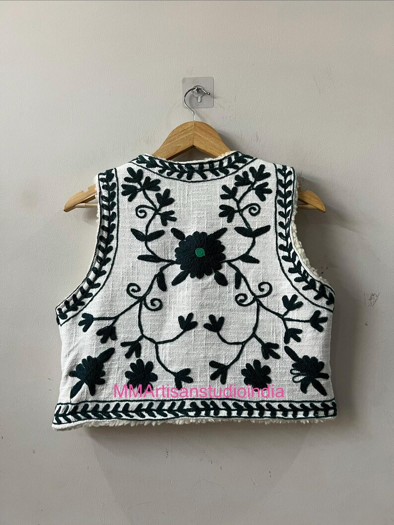 May include: White vest with intricate dark green floral embroidery. The sleeveless garment features a cream-colored sherpa lining and is displayed on a wooden hanger. The vest's design includes a central flower and leafy vines.