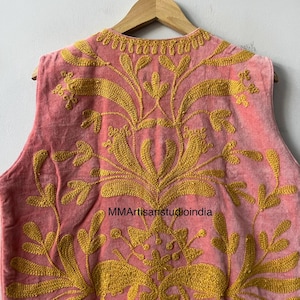 May include: A pink vest with intricate gold embroidery. The vest features a floral design and is hanging on a wooden hanger. The text "MMArtisanstudioindia" is visible on the vest.