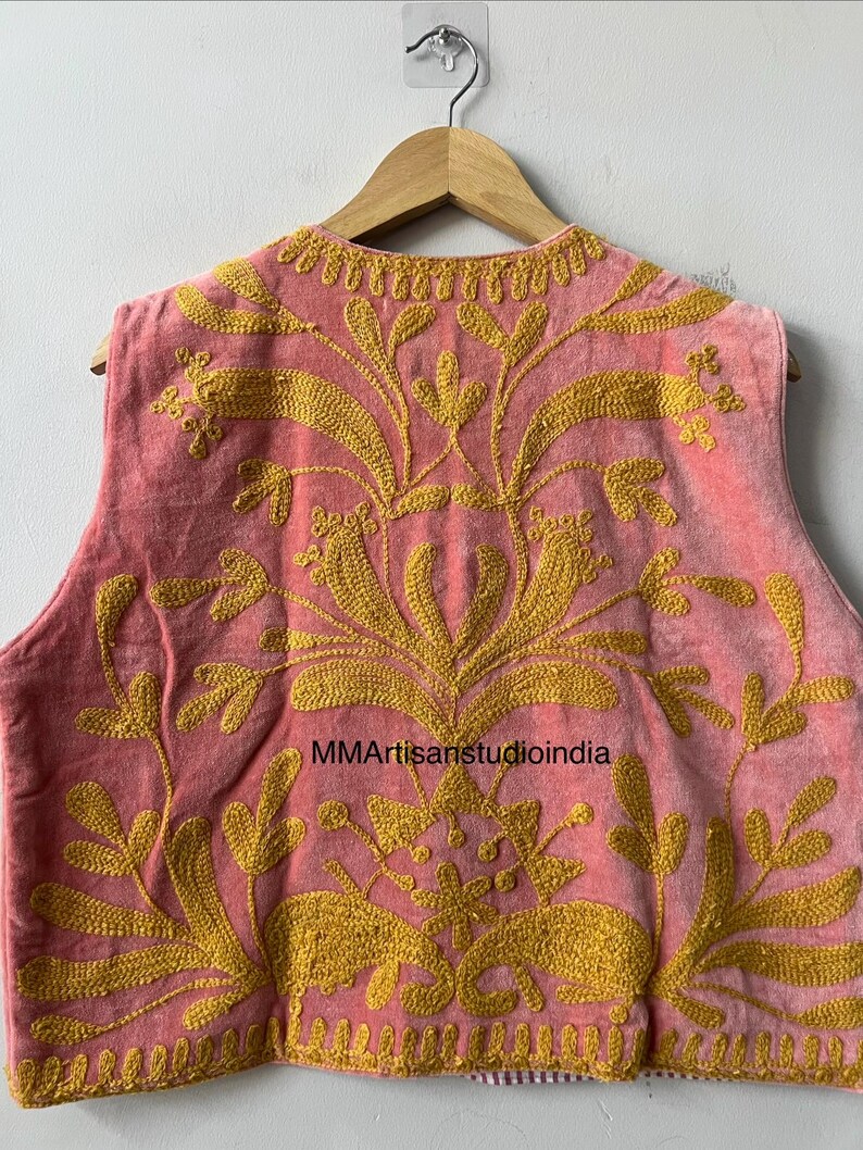 May include: A pink vest with intricate gold embroidery. The vest features a floral design and is hanging on a wooden hanger. The text "MMArtisanstudioindia" is visible on the vest.