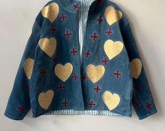 Könnte beinhalten: Eine tealblaue Jacke mit goldenen Herzapplikationen und roten Kreuzakzenten. Die Jacke hat ein gestreiftes Futter und eine lockere Passform. Sie hängt an einem Metallbügel vor einer weißen Wand. Ein einzigartiges Kleidungsstück.