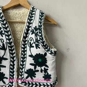 May include: White vest with dark green floral embroidery and a cream-colored sherpa lining. The vest is hanging on a wooden hanger. The embroidery includes floral and leaf patterns.