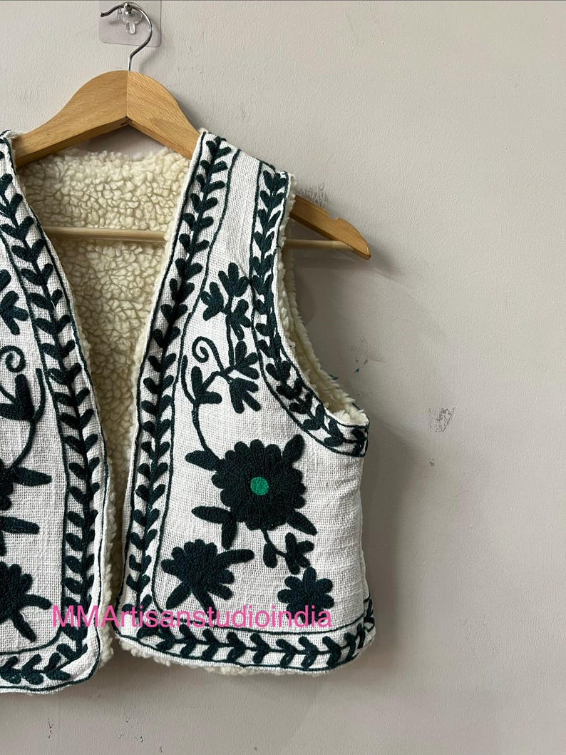 May include: White vest with dark green floral embroidery and a cream-colored sherpa lining. The vest is hanging on a wooden hanger. The embroidery includes floral and leaf patterns.