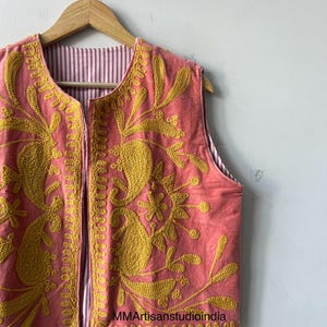 May include: A sleeveless, coral-colored vest with intricate yellow floral embroidery. The vest is reversible, with a striped pink and white lining. The garment is hanging on a wooden hanger against a white wall.