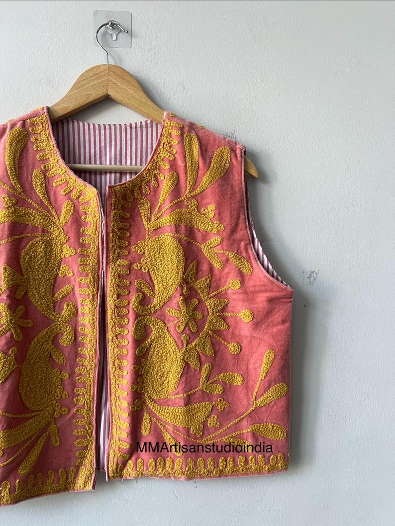 May include: A sleeveless, coral-colored vest with intricate yellow floral embroidery. The vest is reversible, with a striped pink and white lining. The garment is hanging on a wooden hanger against a white wall.